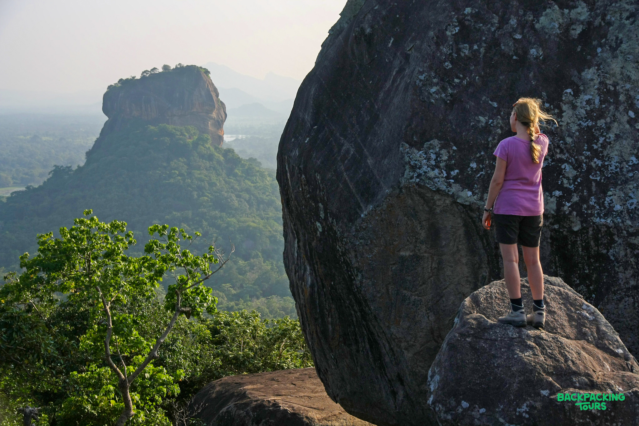 Hiker stands on a rock, gazing at the iconic Sigiriya Lion Rock in Sri Lanka's lush landscape.