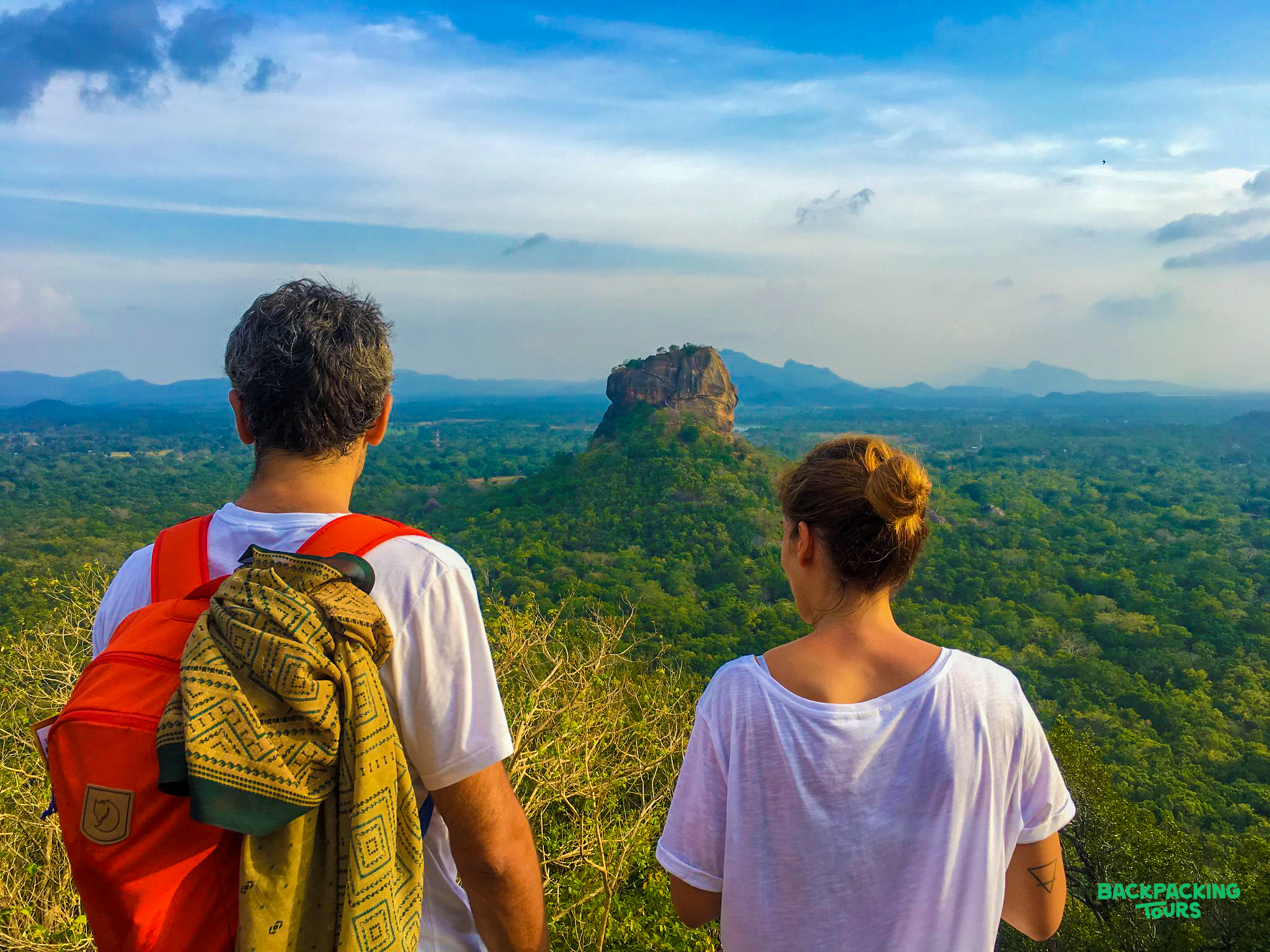 Two travelers gaze at the majestic Sigiriya Lion Rock, surrounded by lush green landscape.