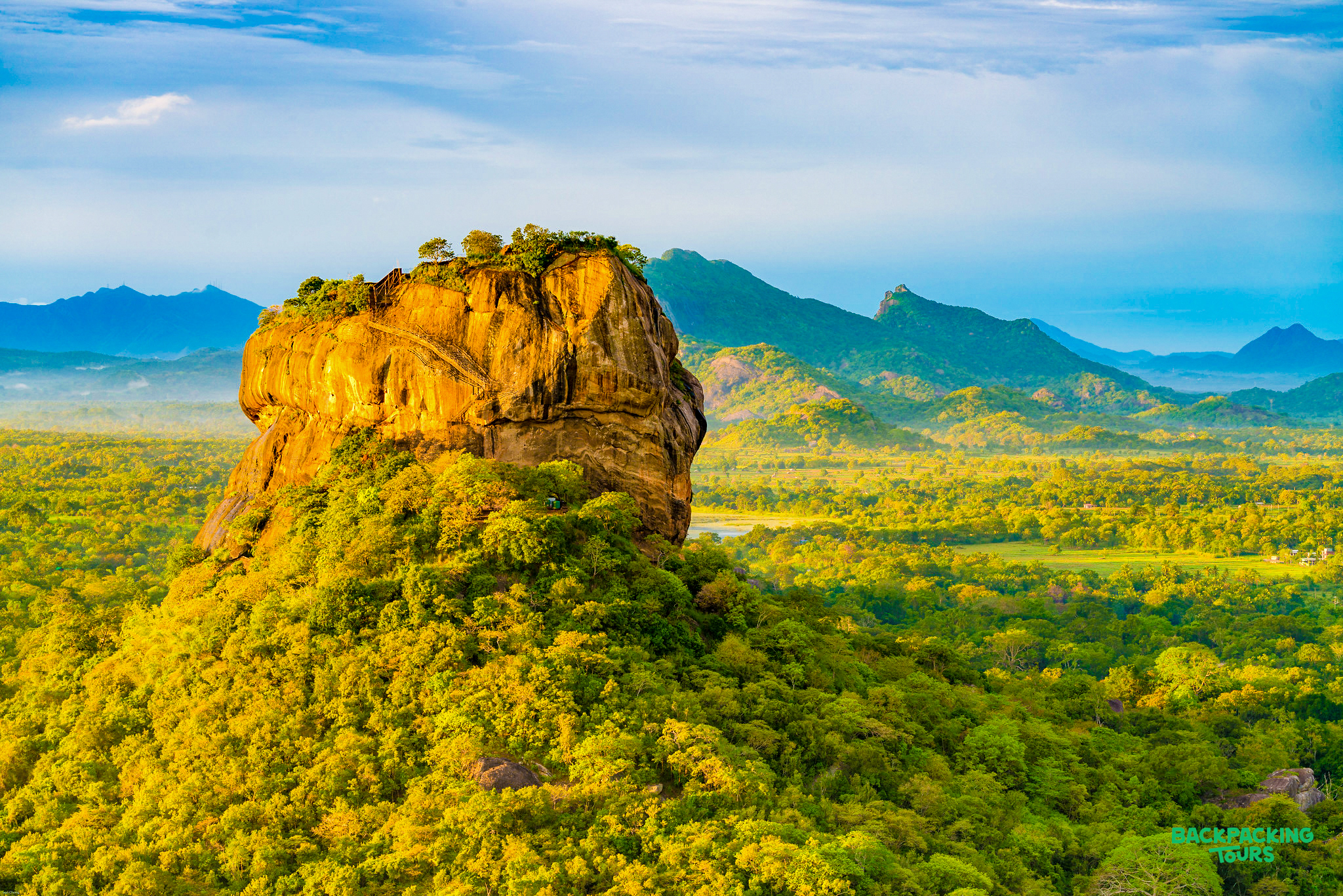 The majestic Sigiriya Lion Rock rises above a dense green forest under a bright blue sky.