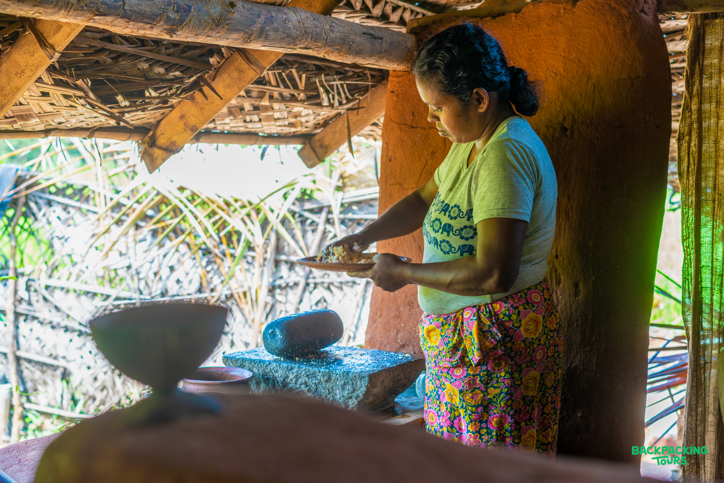 A local woman prepares a traditional meal using a stone grinder in a rustic Sri Lankan kitchen.