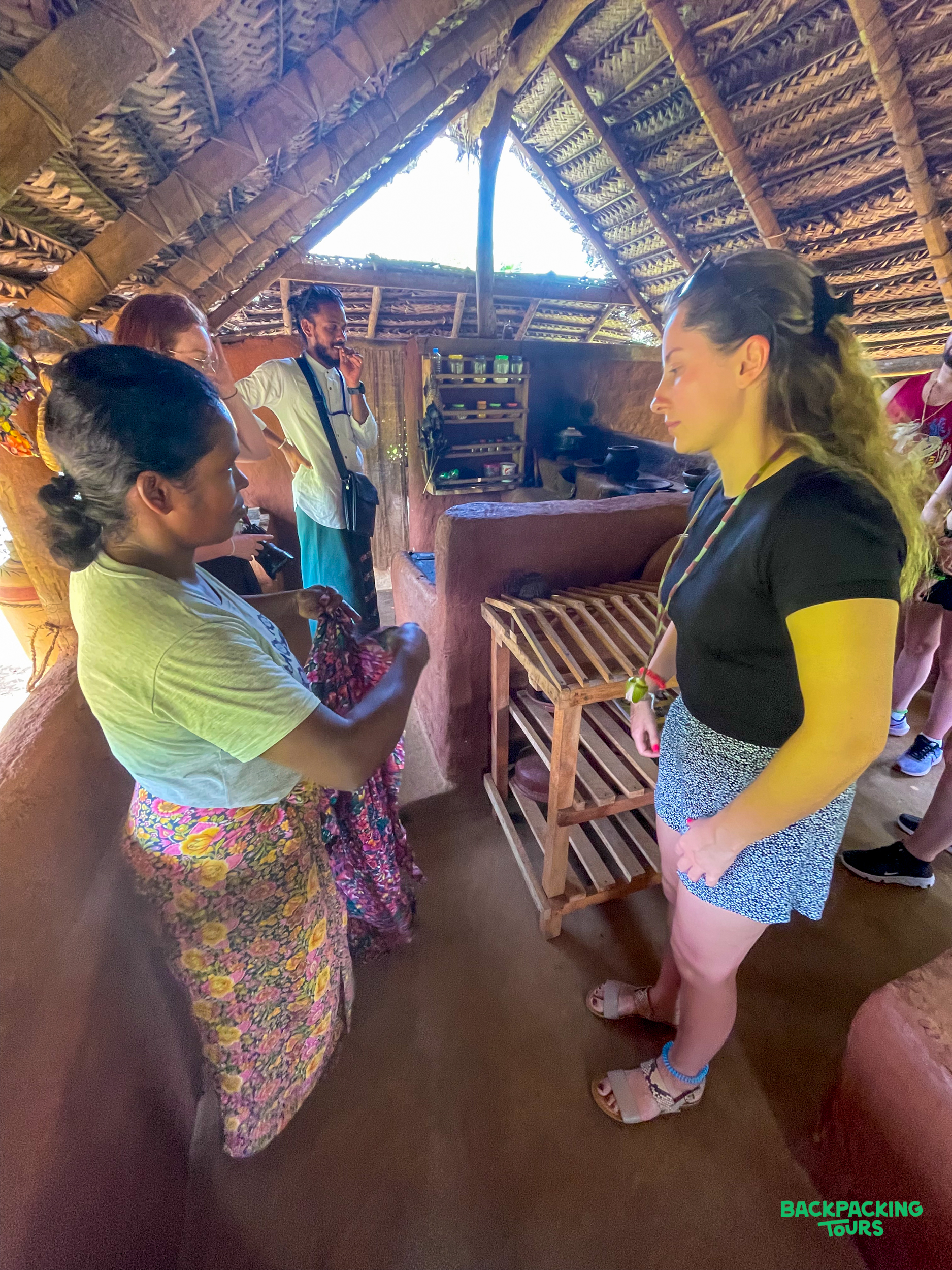 Travelers engage with a local woman showing traditional fabric inside a thatched hut in Kandy.