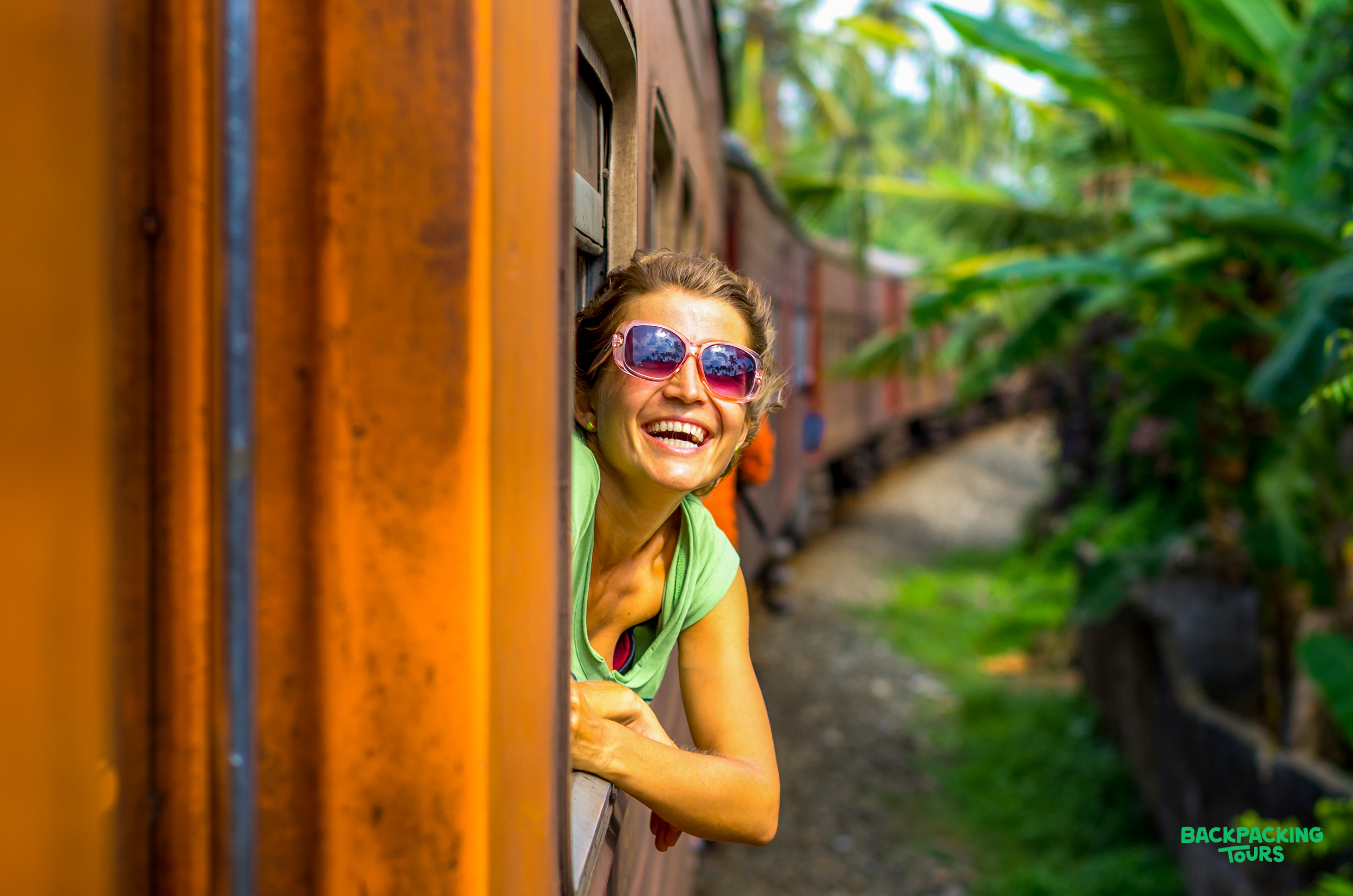 A joyful woman in sunglasses leans out a train window, enjoying the scenic Sri Lankan journey.