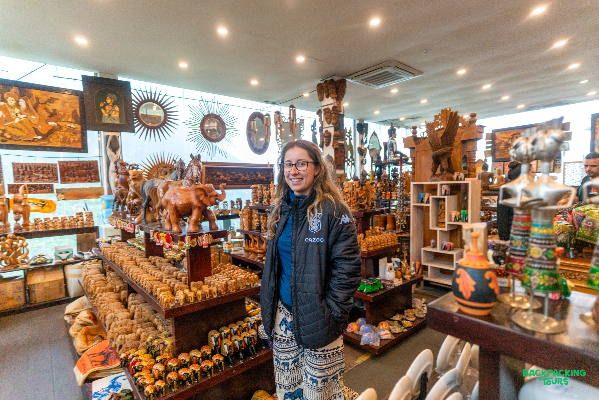 A smiling woman stands in a Sri Lankan souvenir shop filled with carved wooden elephants and local crafts.
