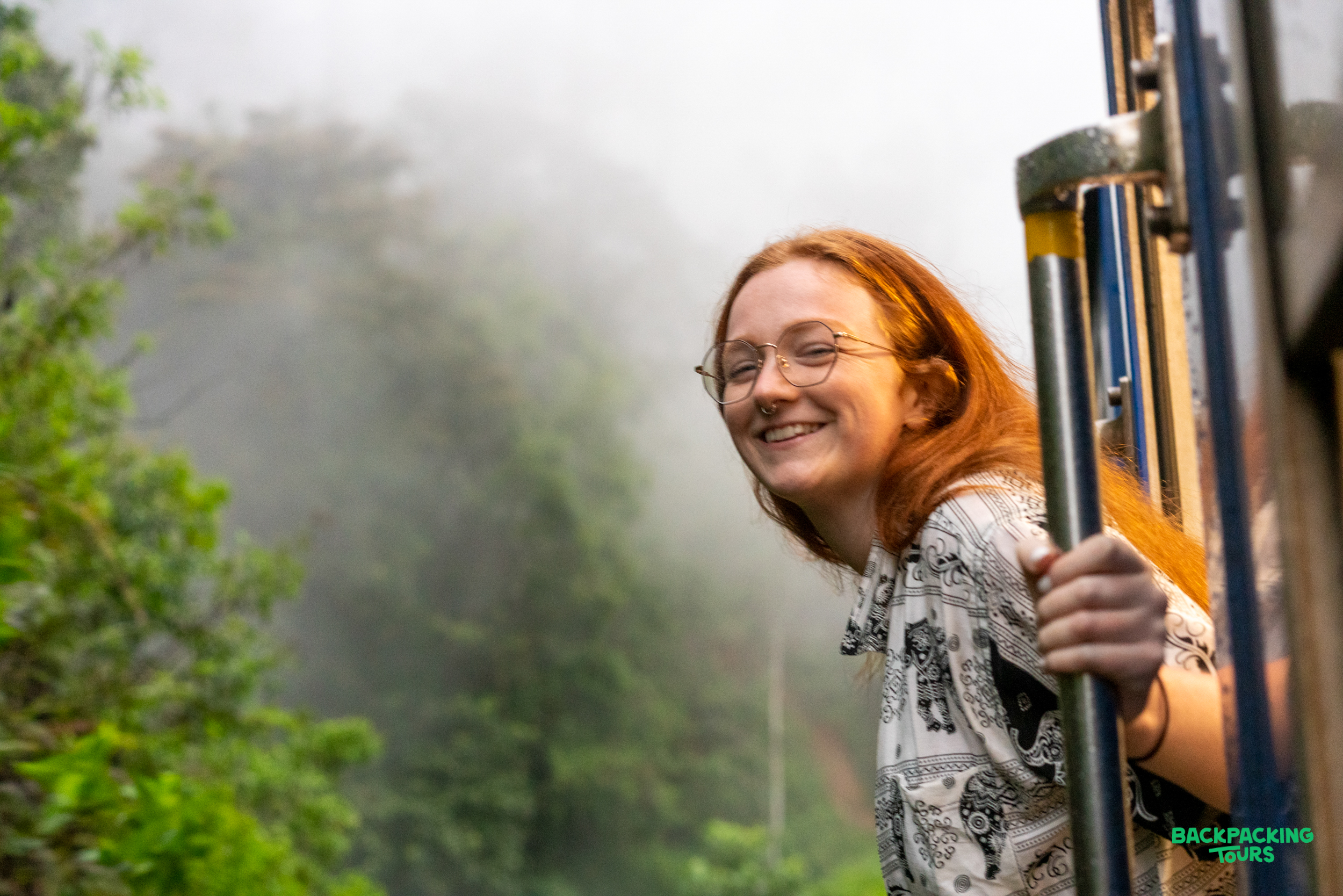 A smiling woman with red hair enjoys the famous train ride through misty Sri Lankan hills.