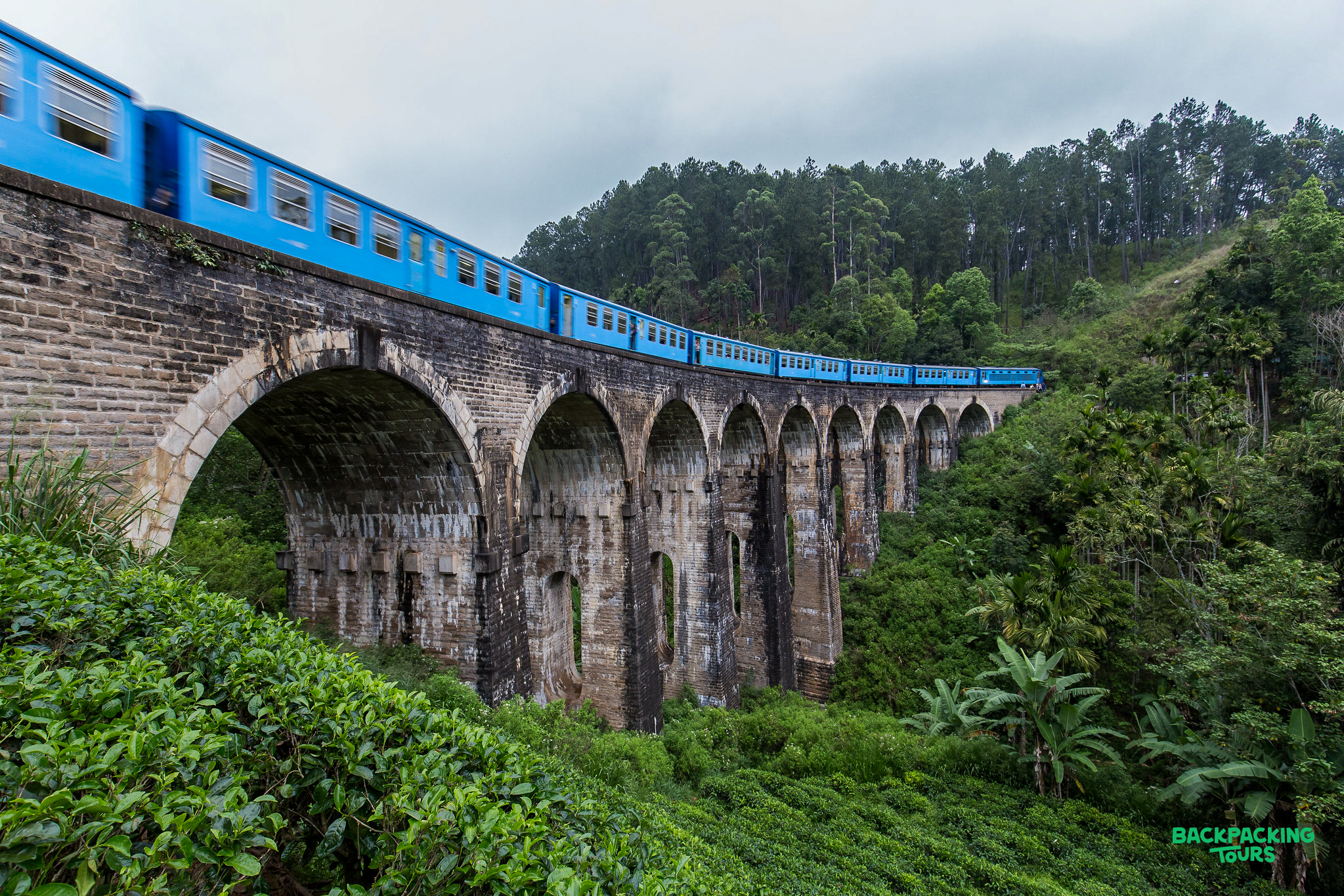 A blue train crosses the historic Nine Arch Bridge, surrounded by lush green tea plantations in Ella, Sri Lanka.