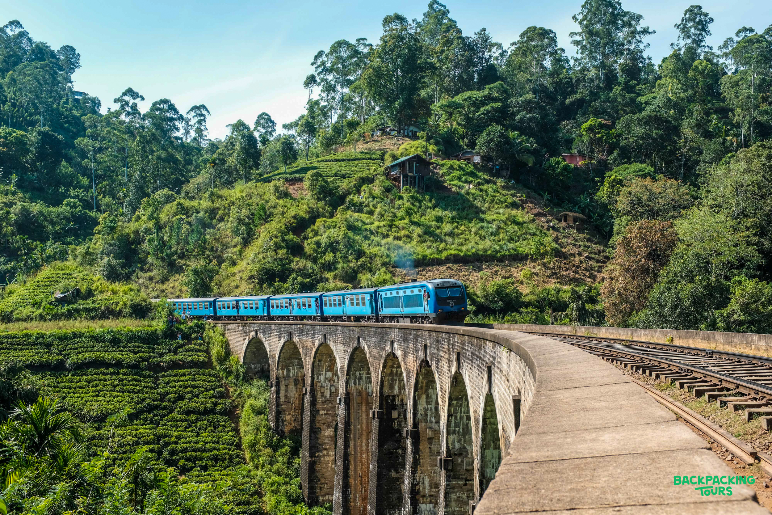 A blue train crosses a historic stone bridge over lush green tea plantations in Sri Lanka.