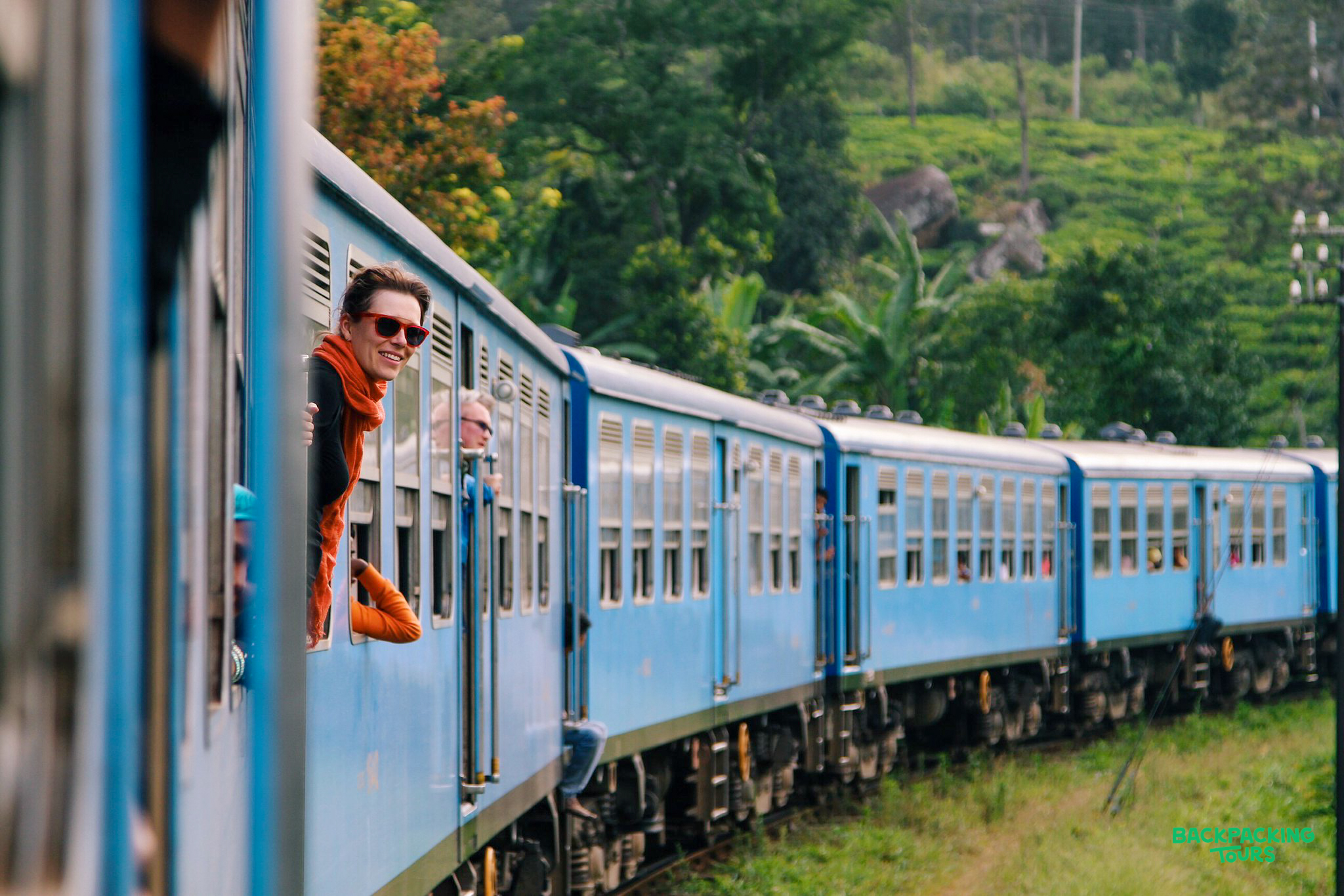 A smiling traveler leans from a blue train window, enjoying the scenic journey through Sri Lanka's lush hills.