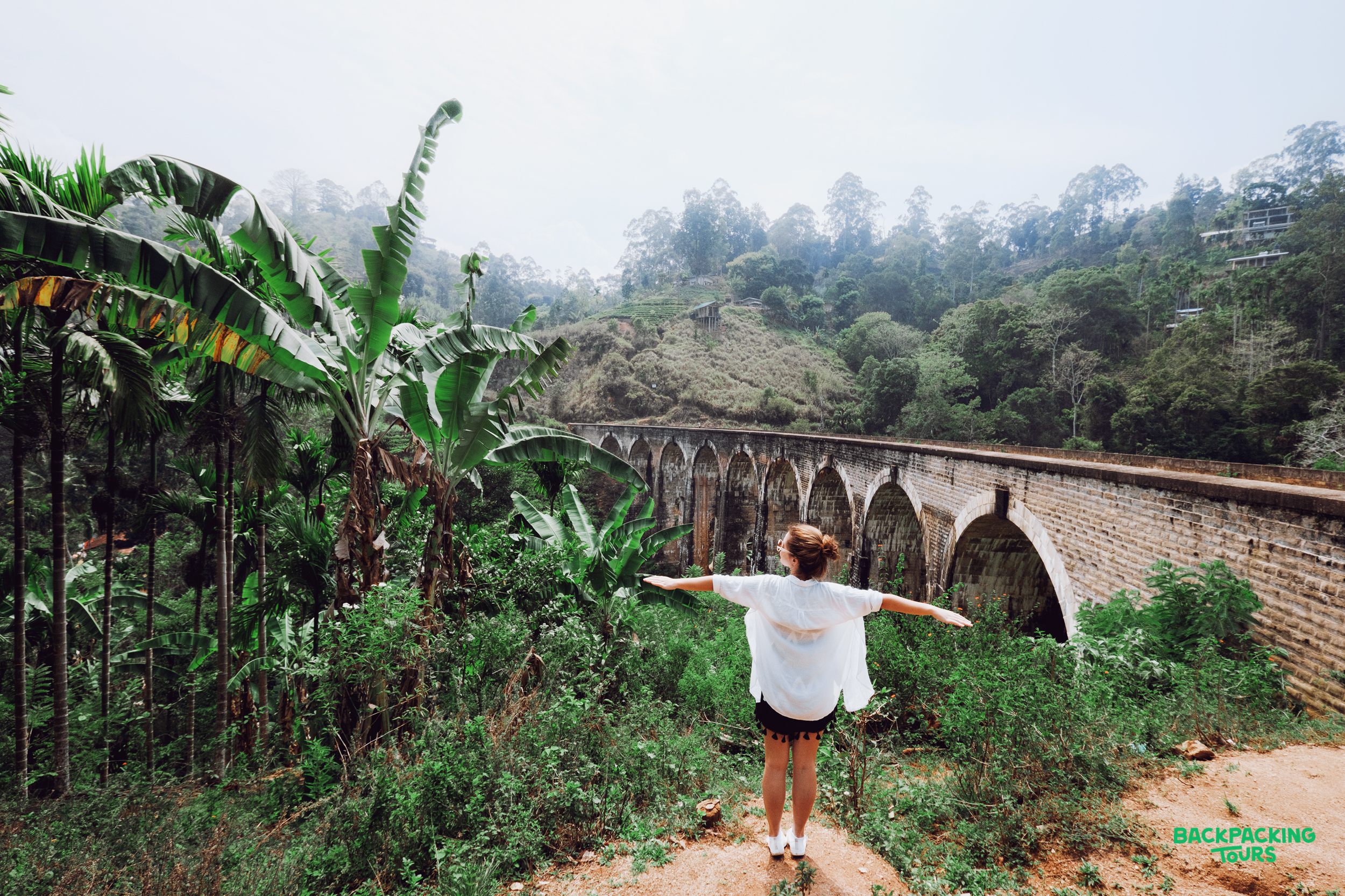A traveler stands with arms outstretched, admiring the iconic Nine Arch Bridge surrounded by lush greenery in Ella, Sri Lanka.