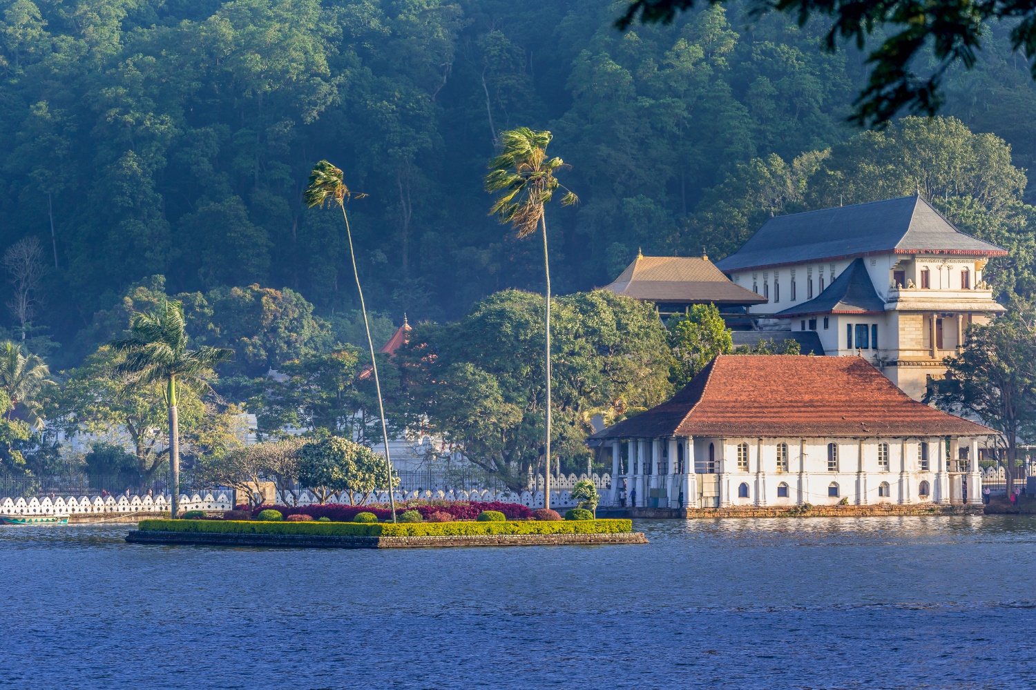 Historic Kandy temple complex by a serene lake with palm trees and lush green hills.