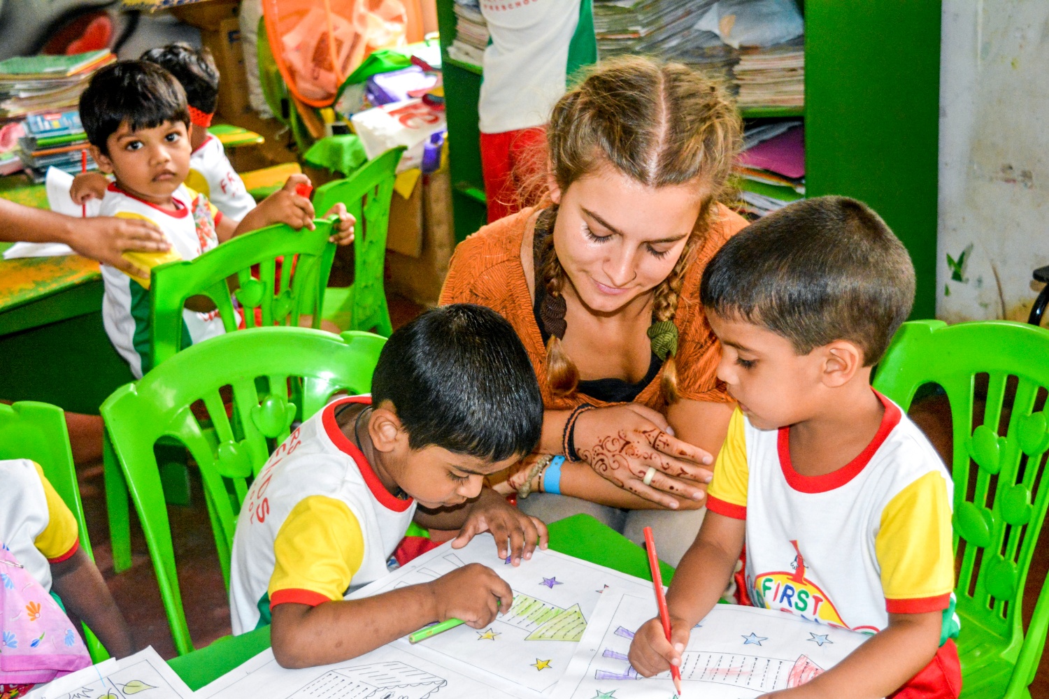 A female volunteer helps two young Sri Lankan boys color drawings in a bright classroom.