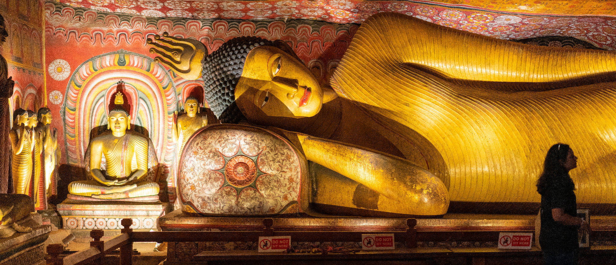 A person views a large golden reclining Buddha statue inside the richly decorated Dambulla Cave Temple.