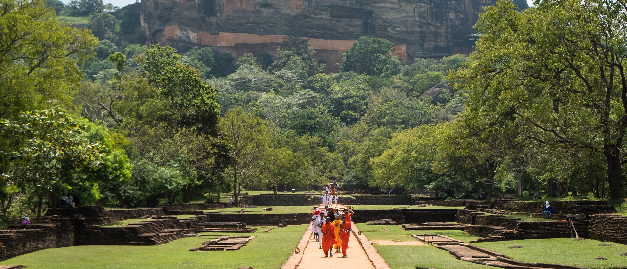 People walk on a path through ancient garden ruins towards the majestic Sigiriya Lion Rock.