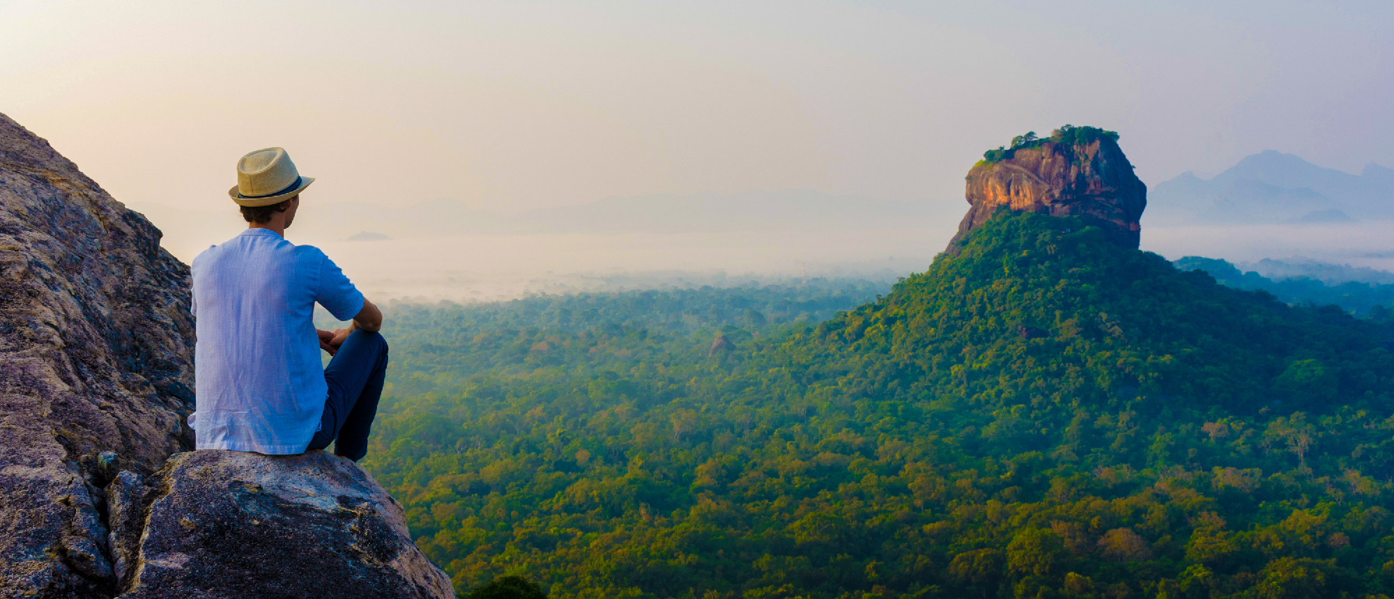 A traveler in a hat sits on a cliff, enjoying panoramic views of Sigiriya Rock and lush forests.