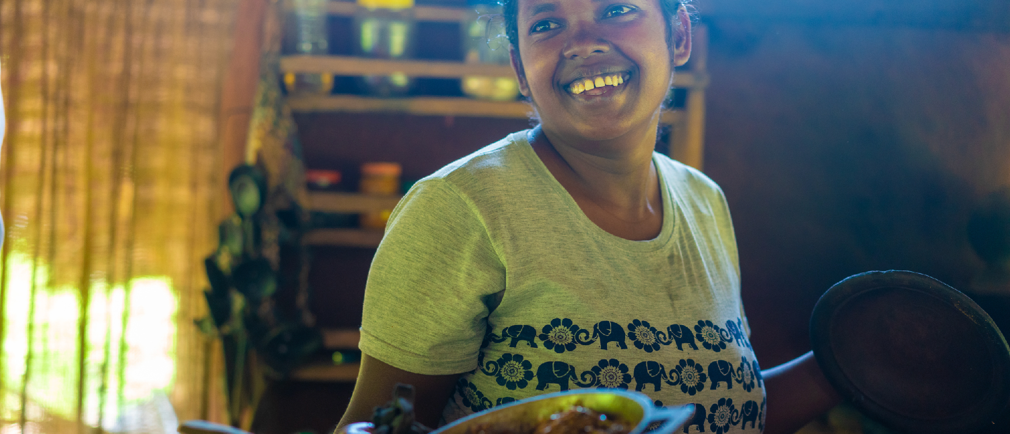 A joyful woman in Sri Lanka holds a pot of traditional food, smiling brightly during a village tour.