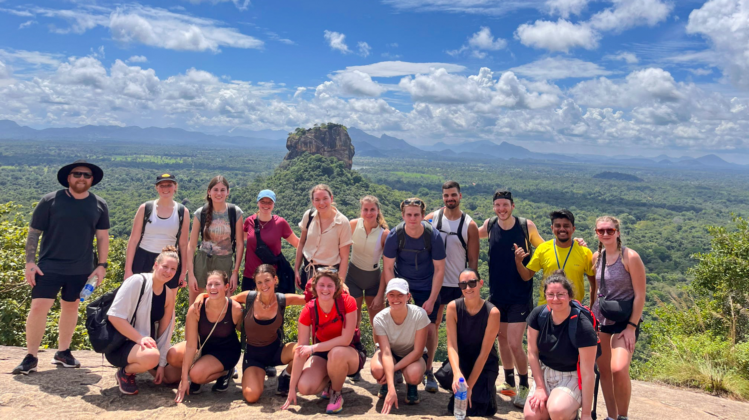 A large group of happy hikers posing on Pidurangala Rock with Sigiriya in the background.