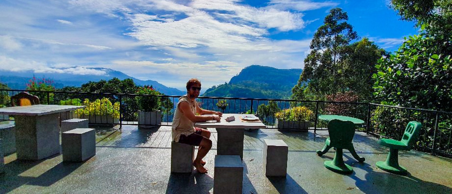 A person relaxes at a table on a terrace, enjoying a panoramic view of Udawalawe's green mountains.