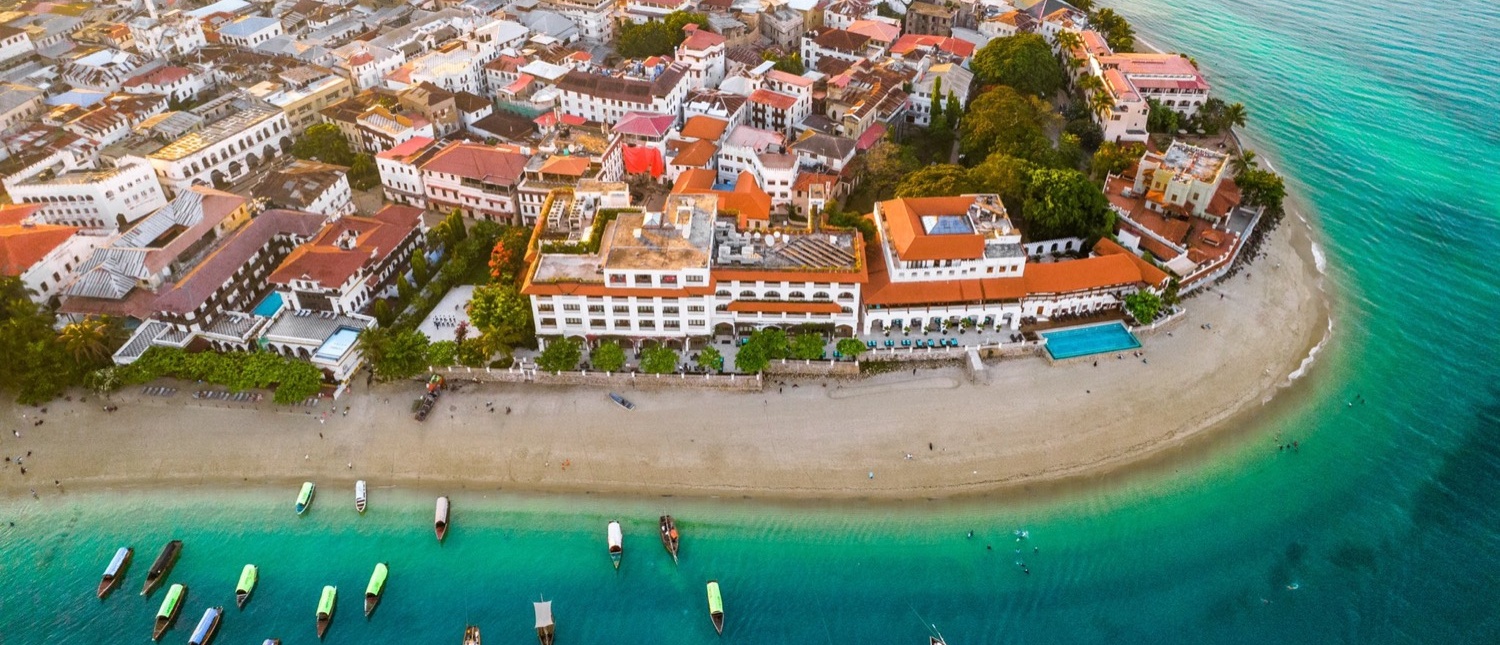 Aerial view of Zanzibar's historic Stone Town, featuring a sandy beach, turquoise ocean, and many boats.