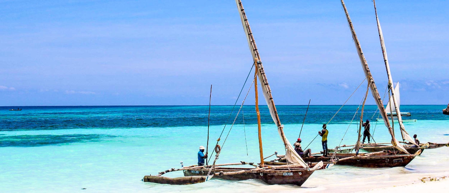 People working on traditional dhow boats on a white sand beach with turquoise water in Zanzibar.