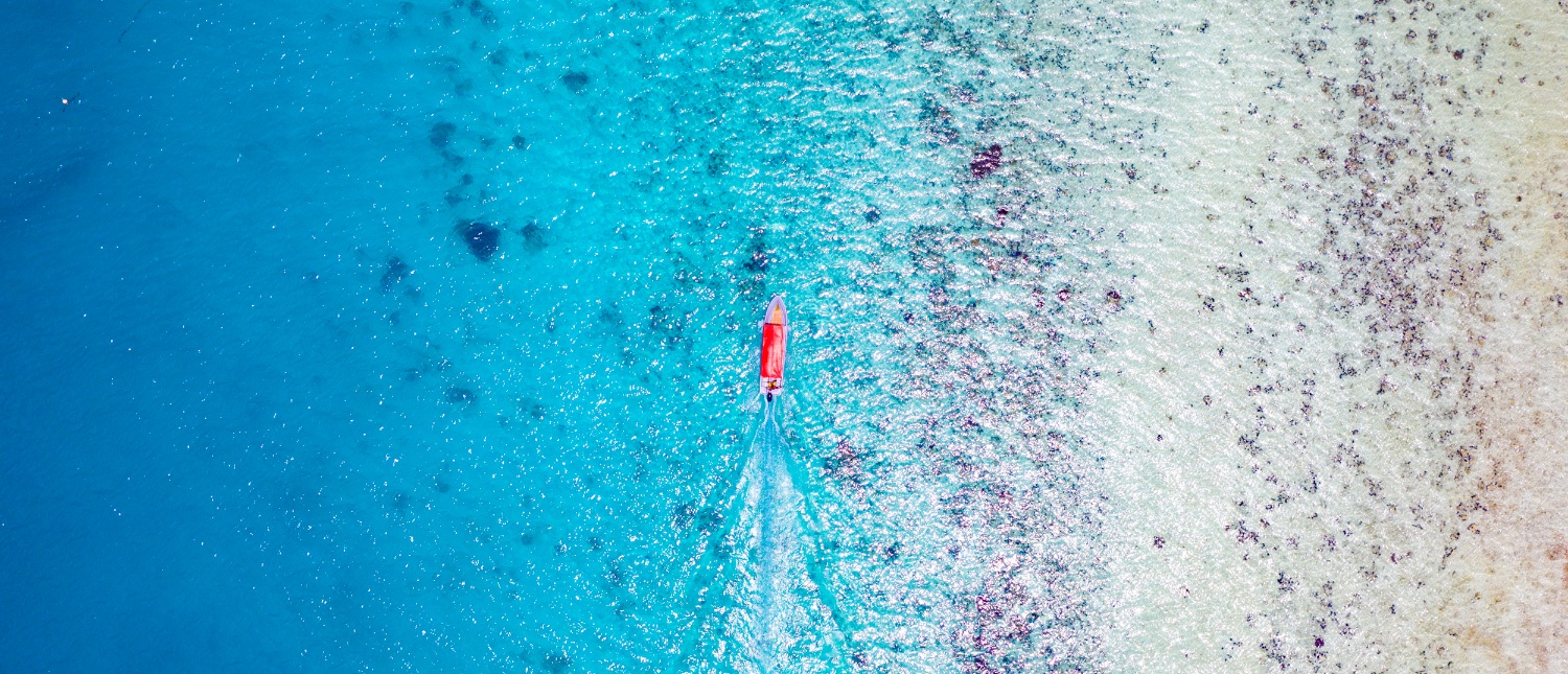An aerial view of a red boat moving through clear turquoise water near a shallow reef in Zanzibar.