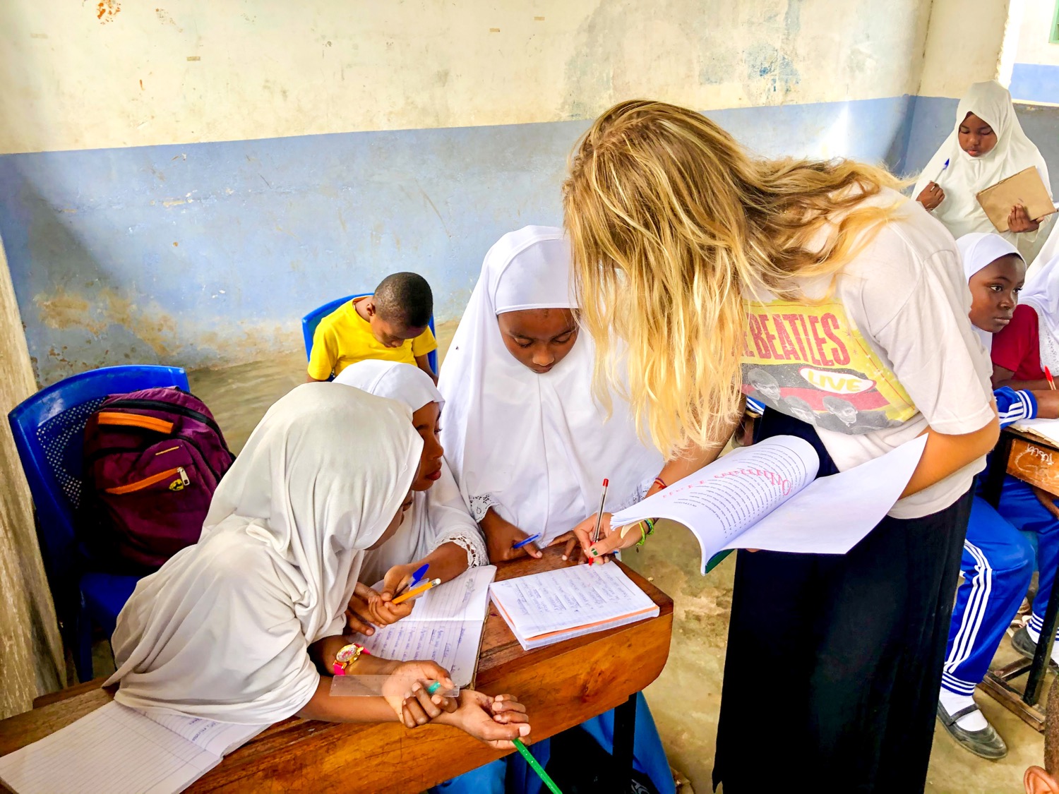 A blonde volunteer teacher helps three young girls in hijabs with their studies in a Zanzibar classroom.