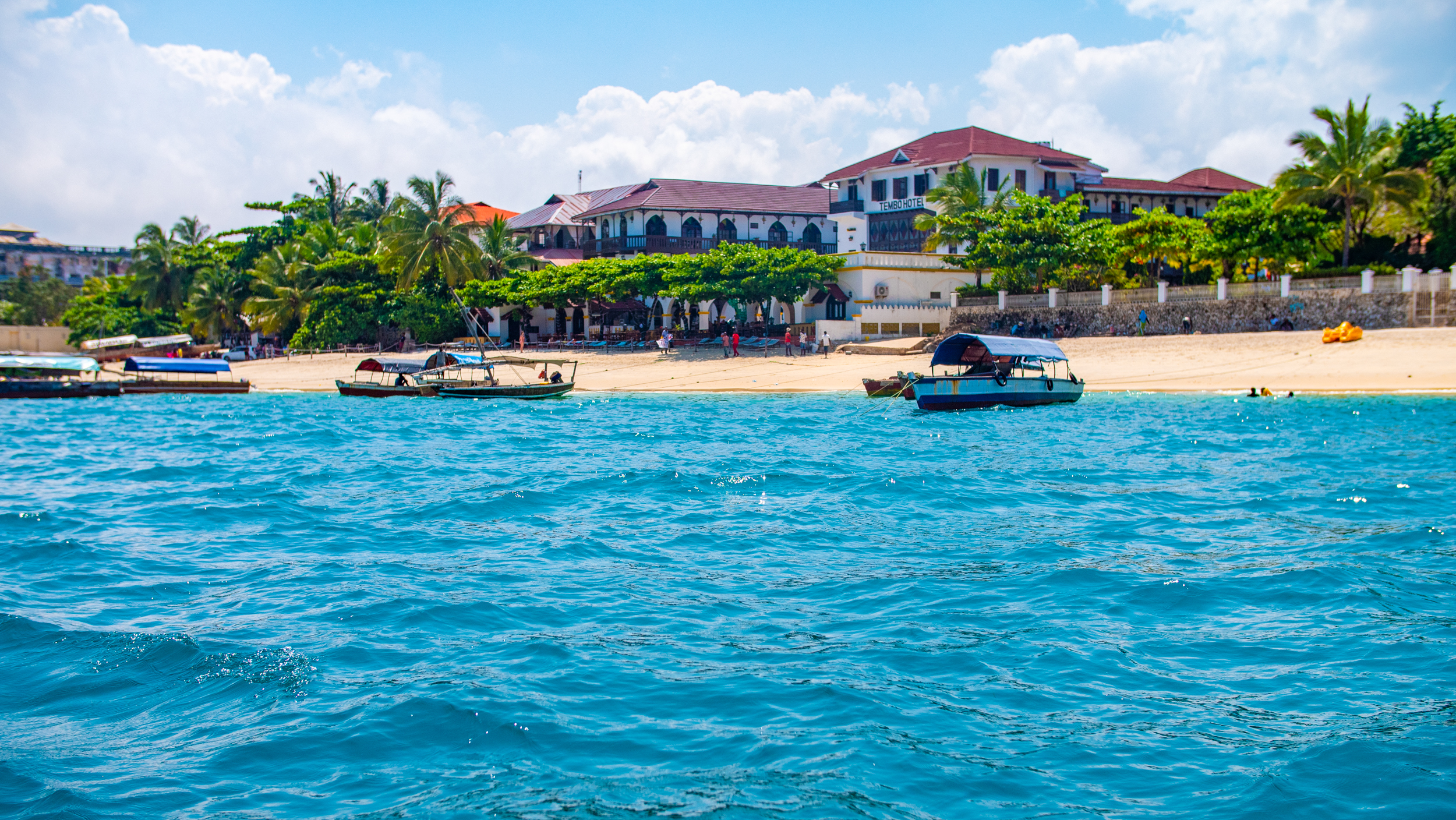 Boats on the clear blue ocean near a sandy beach with large buildings and palm trees in Zanzibar.