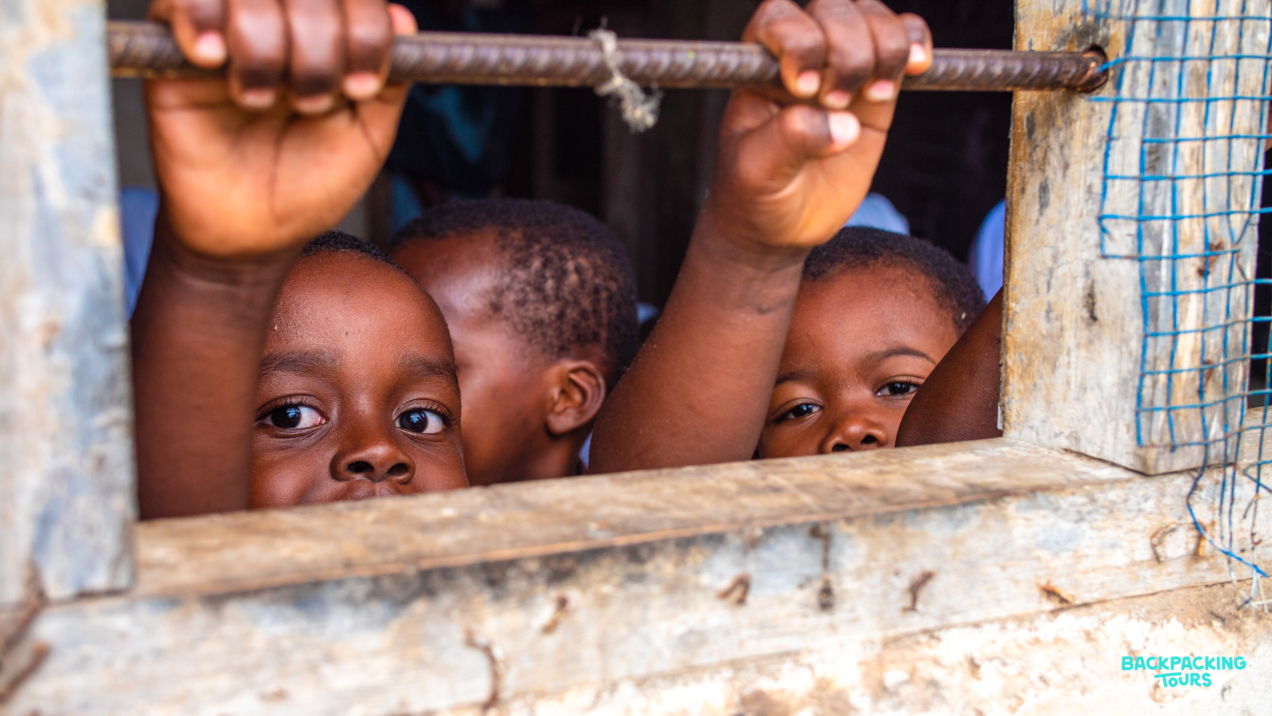 Curious young students in Zanzibar peek out from their classroom window, hands on the bar.