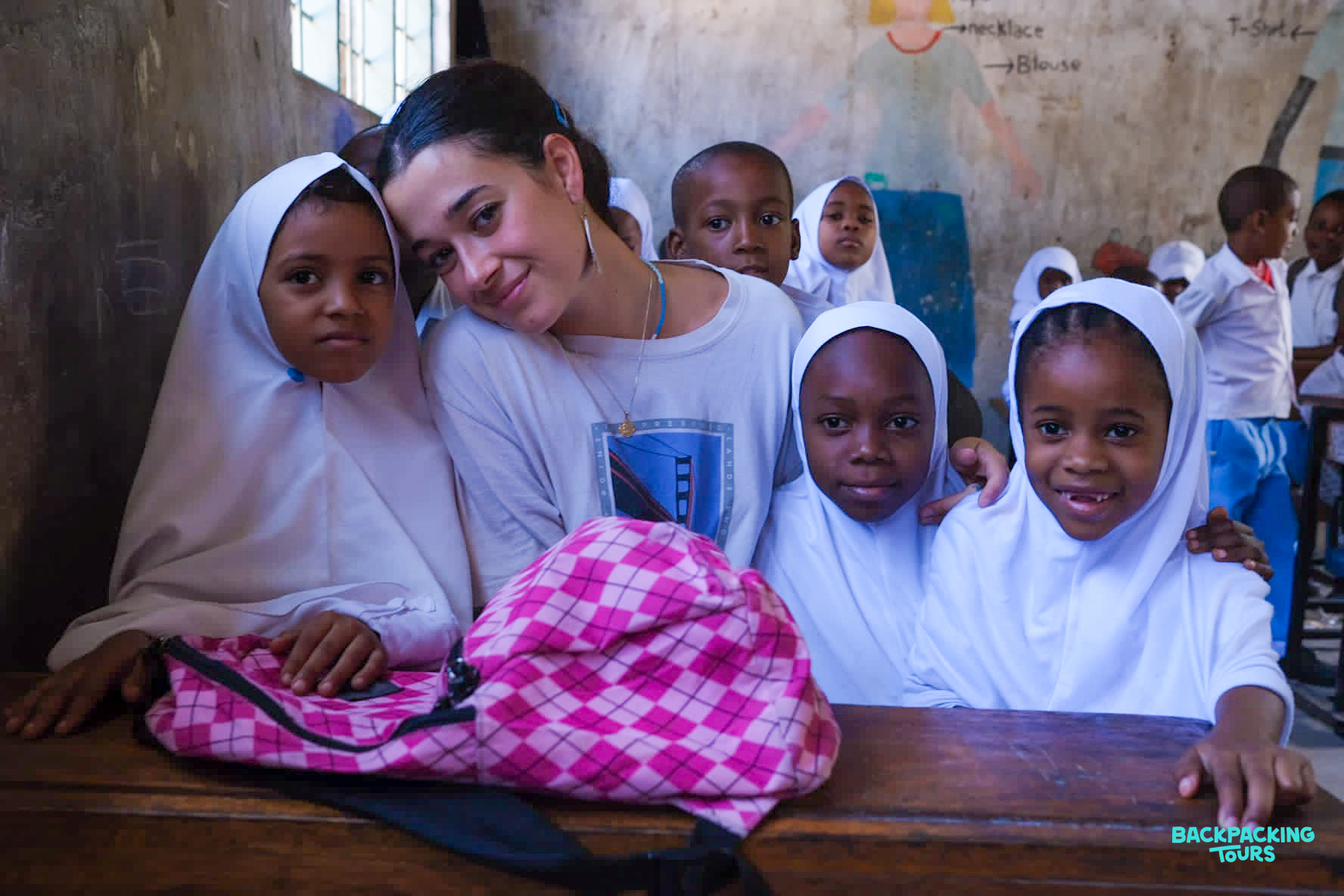 A young volunteer teacher happily poses with smiling students in a classroom in Zanzibar.