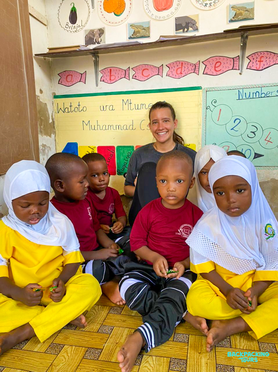 A cheerful volunteer and young children sit on the floor in a vibrant Zanzibar classroom.