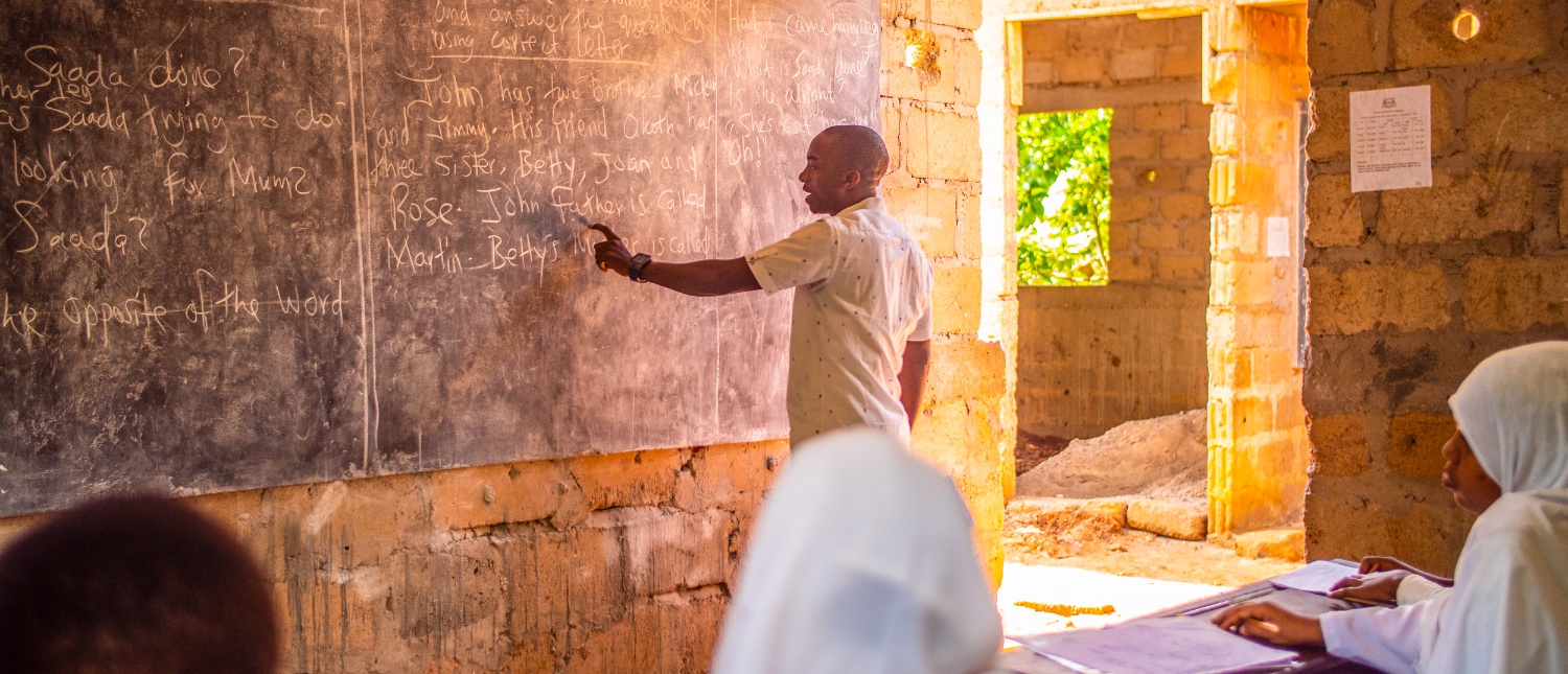 A male teacher points at a blackboard while students listen in a classroom in Zanzibar.
