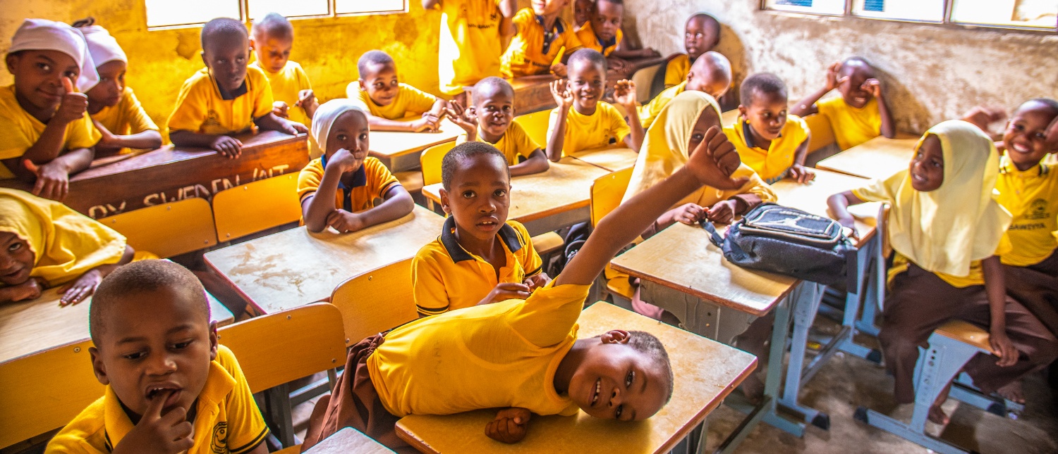 Lively students in yellow uniforms smiling and raising hands in a bright classroom in Zanzibar.