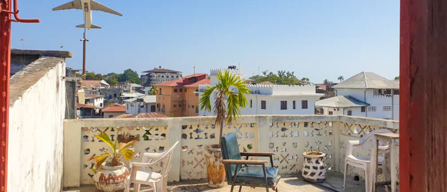 Rooftop terrace in Zanzibar with chairs, potted plants, and a model airplane above city buildings.