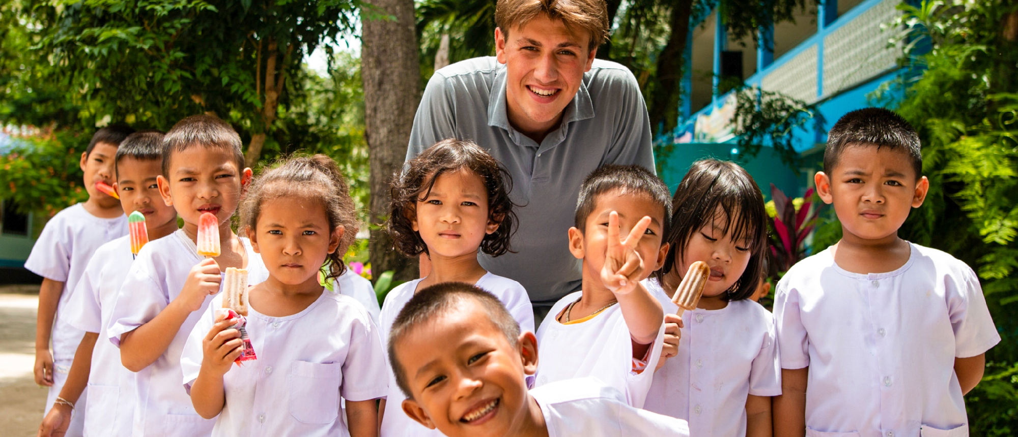 A smiling volunteer with a group of happy Thai children holding popsicles outdoors.