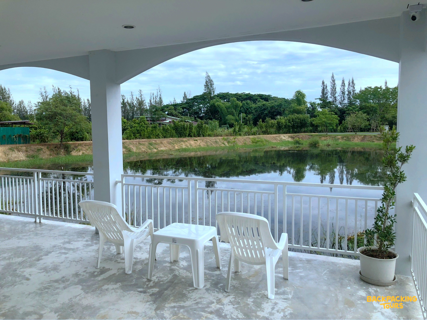 A peaceful outdoor patio with white chairs and a table overlooks a calm lake and lush green trees.