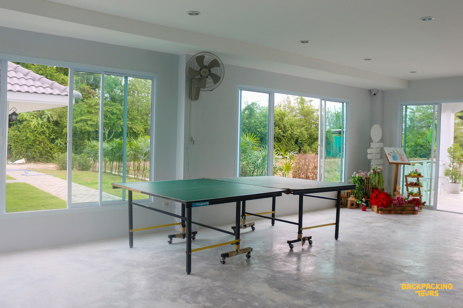Bright common room featuring two ping-pong tables and large windows overlooking a lush garden in Thailand.