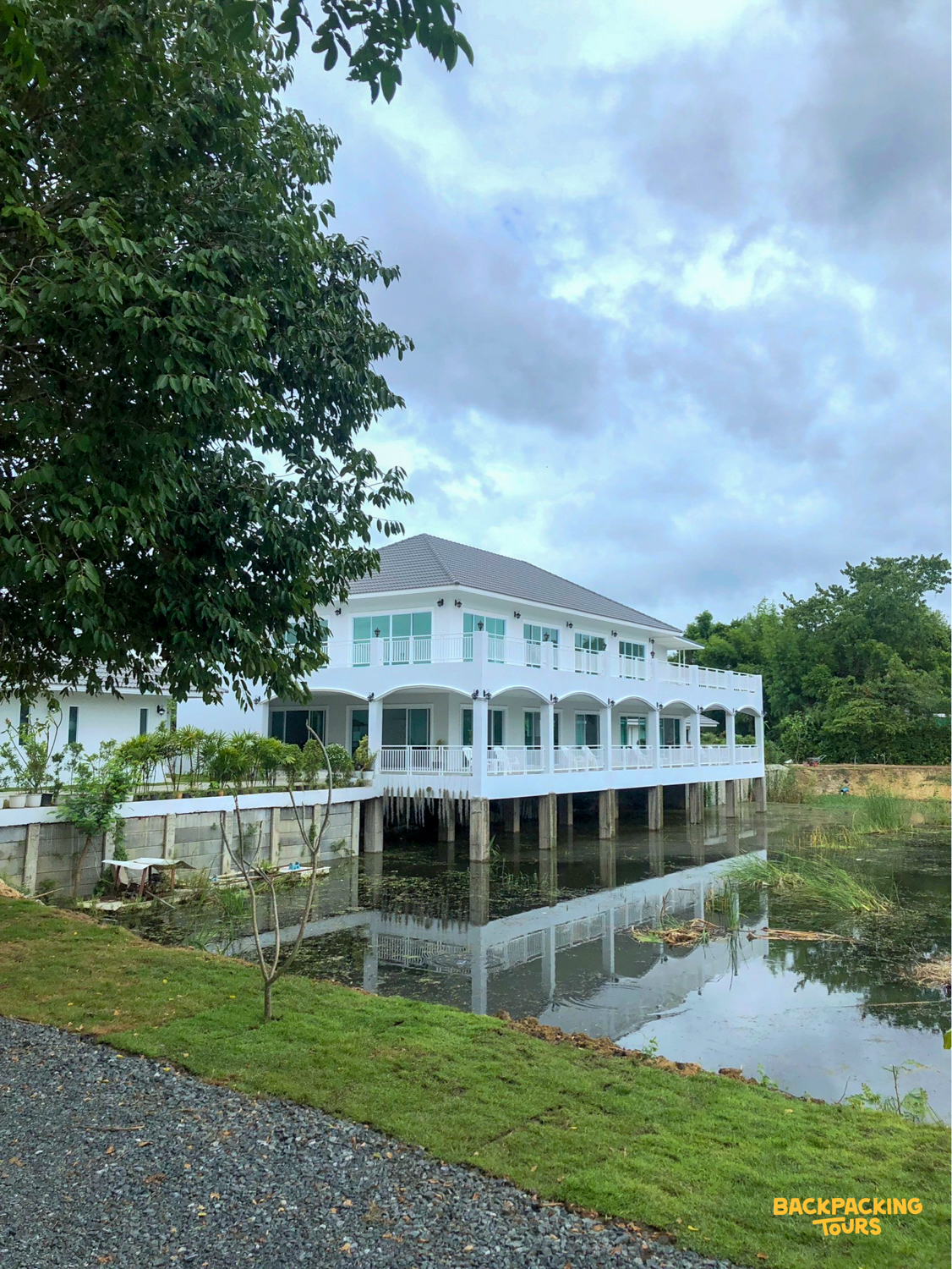 Spacious white accommodation building with balconies overlooking a calm pond under a cloudy sky.