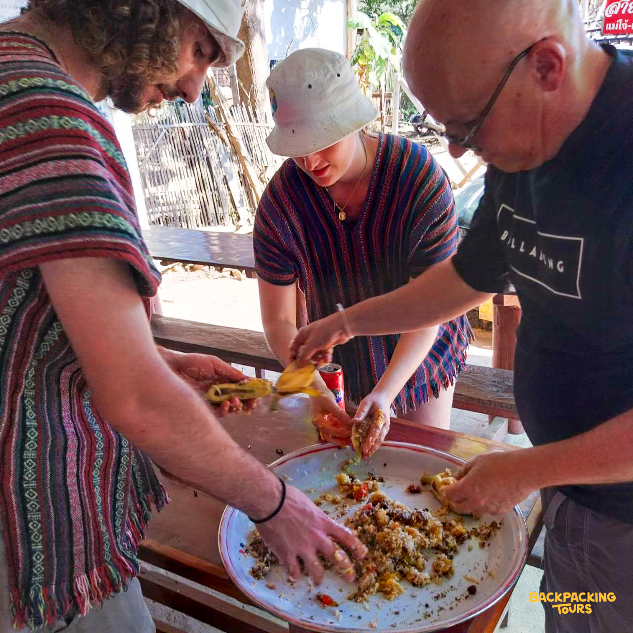 Backpacking volunteers prepare a traditional Thai meal by hand, mixing ingredients in a large tray.