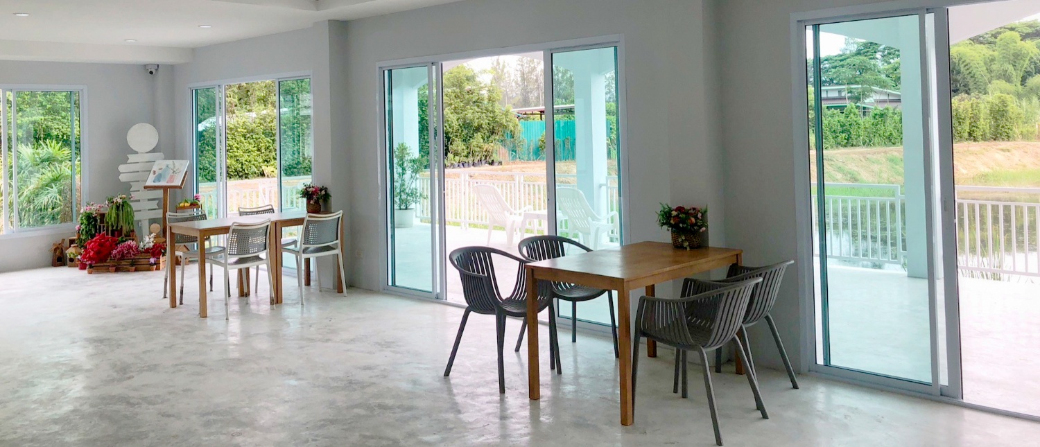 Bright, modern common room with wooden tables and chairs, large windows overlooking lush tropical greenery in Thailand.