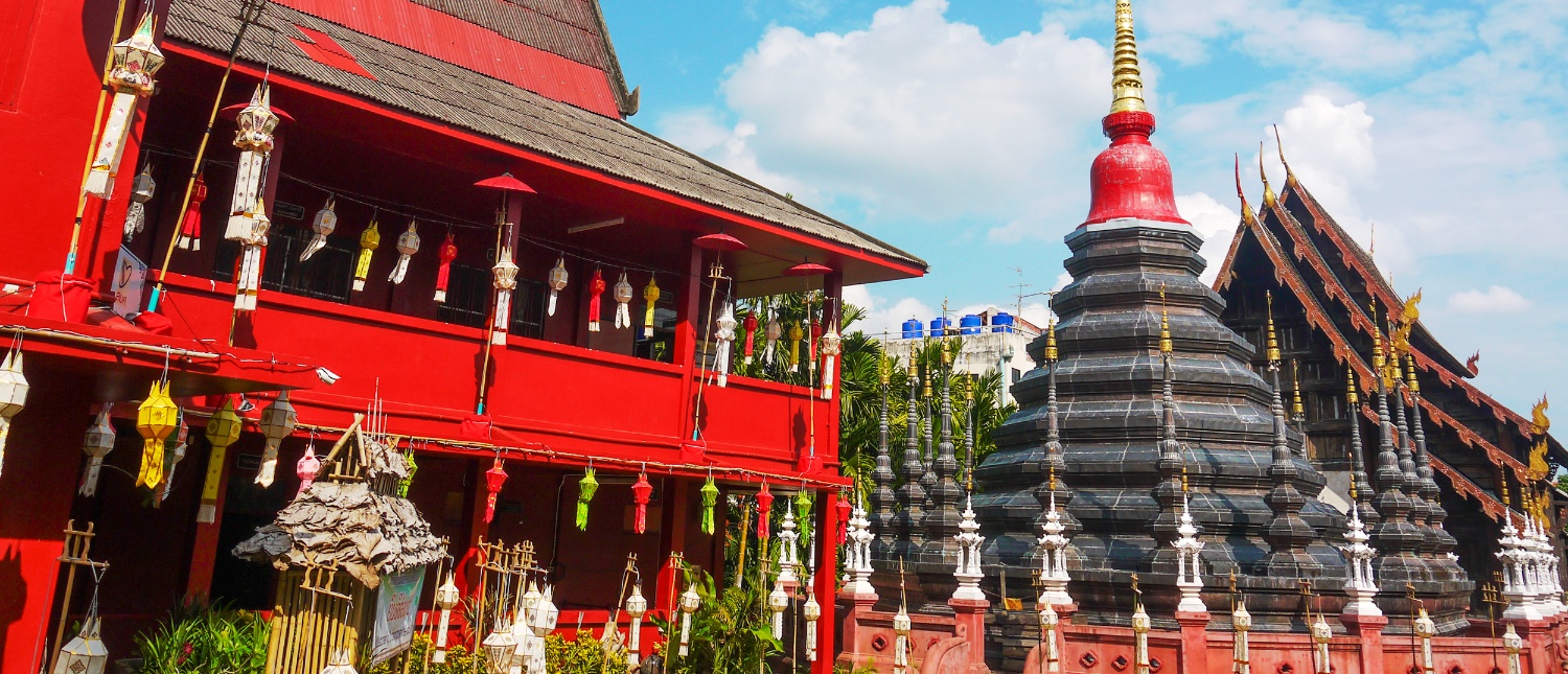 A vibrant red temple building adorned with colorful lanterns next to a tiered grey pagoda in Thailand.