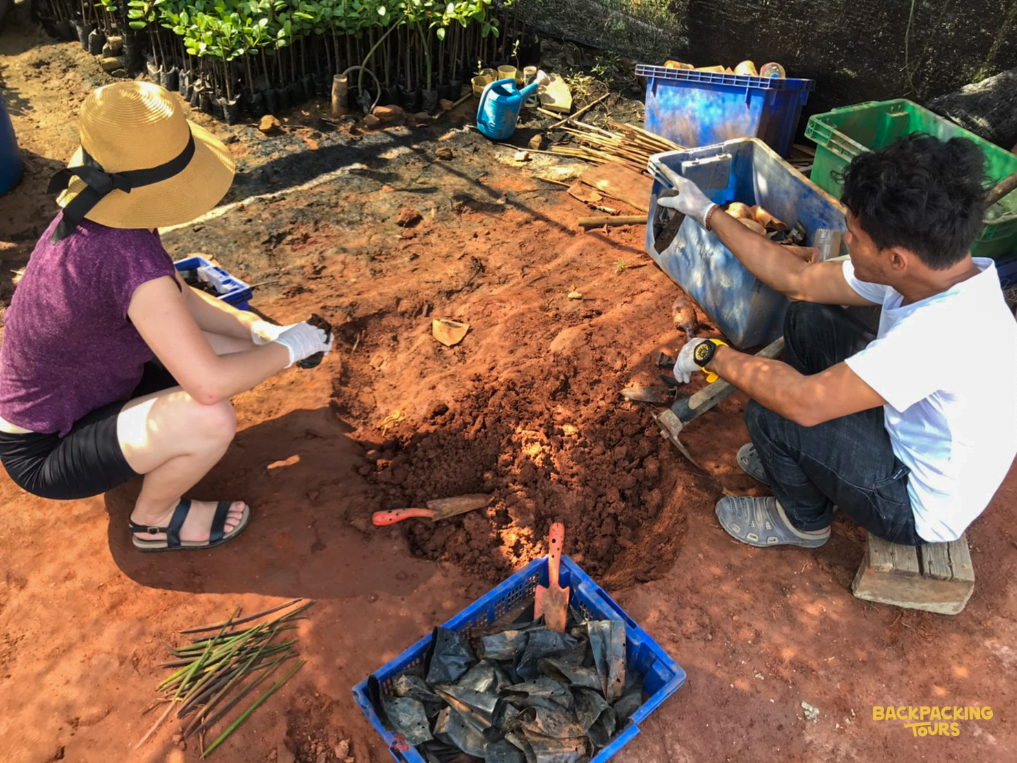 Two volunteers crouch, planting new seedlings into the red soil at a conservation site in Thailand.