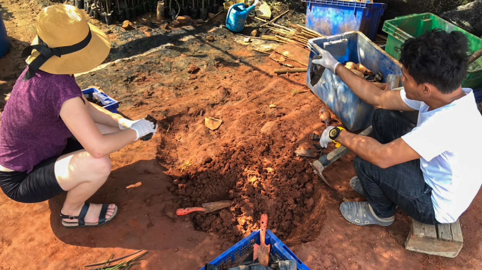 Two volunteers in hats and gloves dig and plant in reddish soil during a conservation project in Thailand.