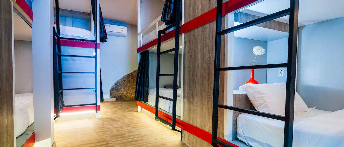 Bright and clean hostel dormitory room featuring modern bunk beds with privacy curtains and red details.