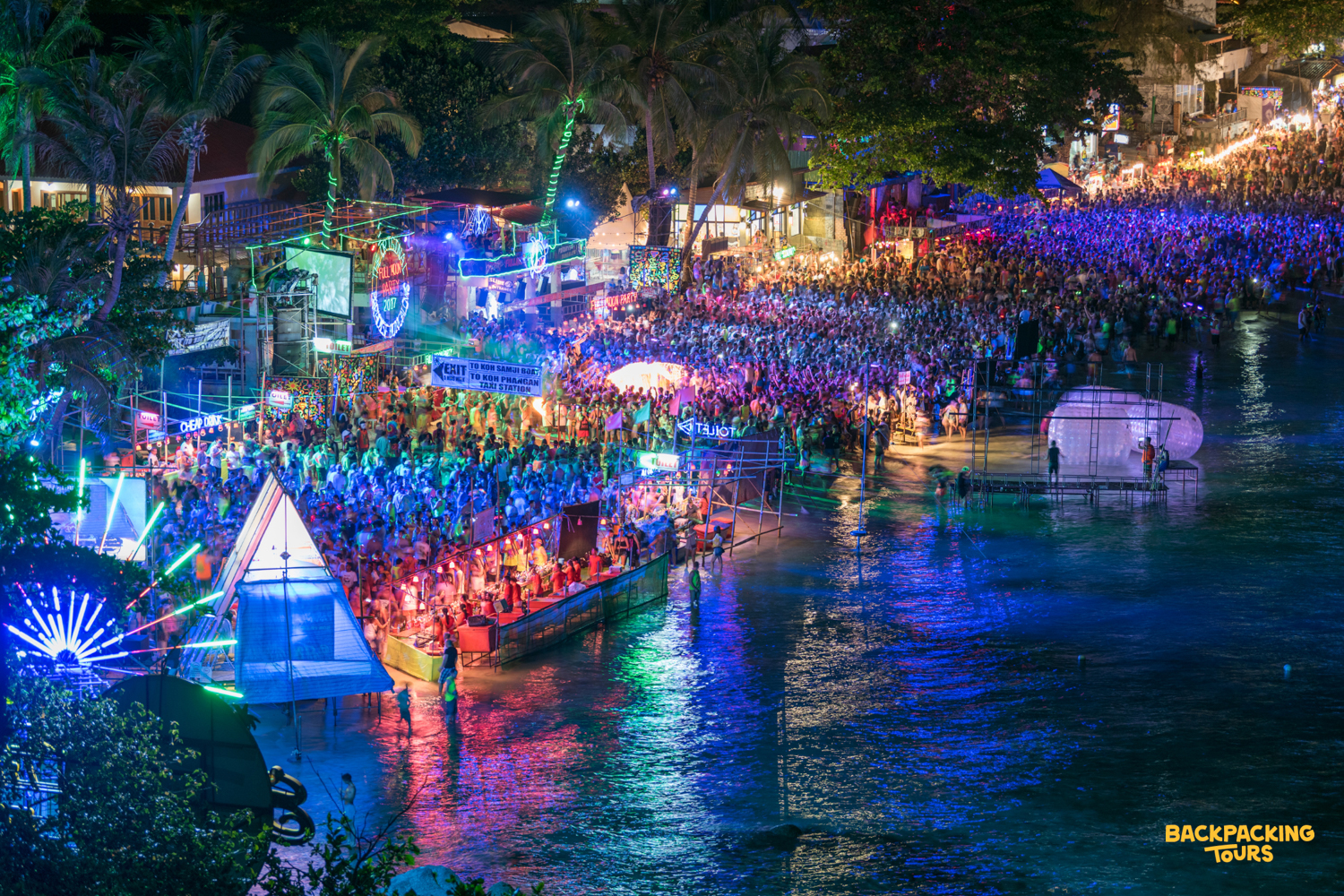 Thousands of people gather for a vibrant beach party on Koh Phangan, illuminated by colorful lights.