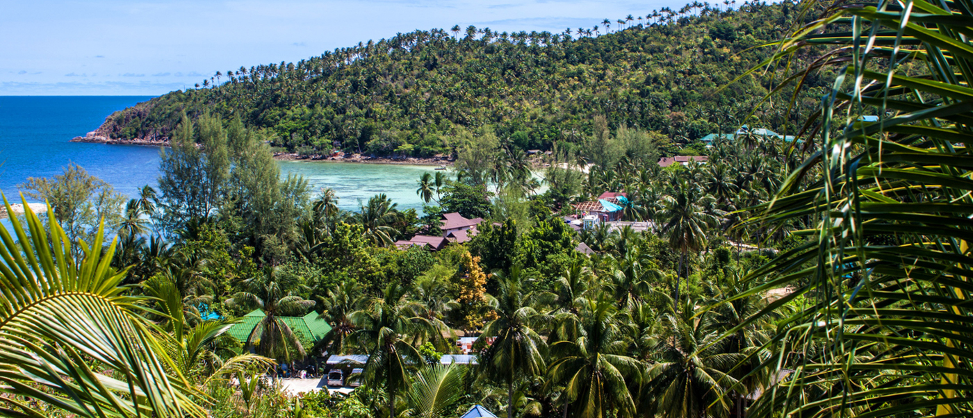 Lush green hills and palm trees overlook a tranquil turquoise bay on Koh Phangan island.