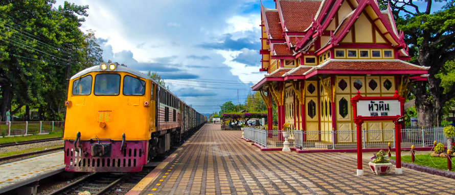 A yellow train waits at the ornate Hua Hin railway station under a cloudy sky.