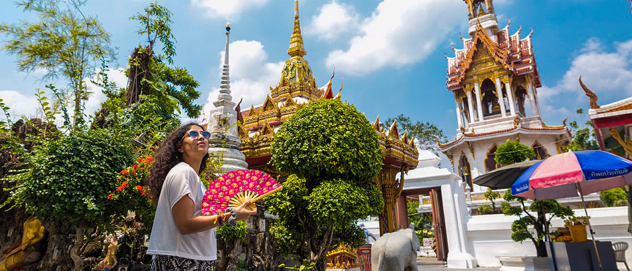A woman with a fan admires the ornate golden temples and white stupas under a sunny Bangkok sky.