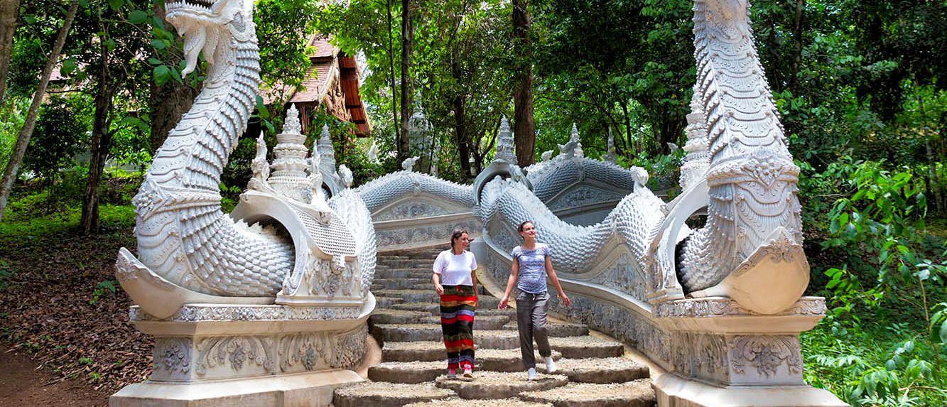 Two women ascend a grand stone staircase flanked by ornate white naga sculptures at a temple in Chiang Mai.