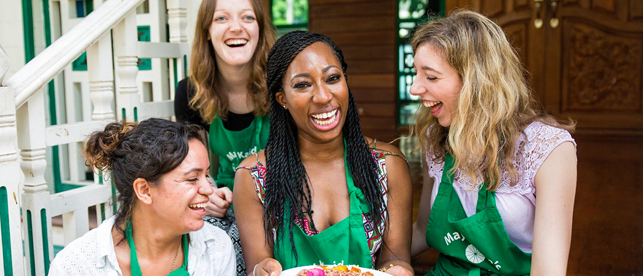 Smiling women in green aprons laugh together during a hands-on cooking class in Bangkok.