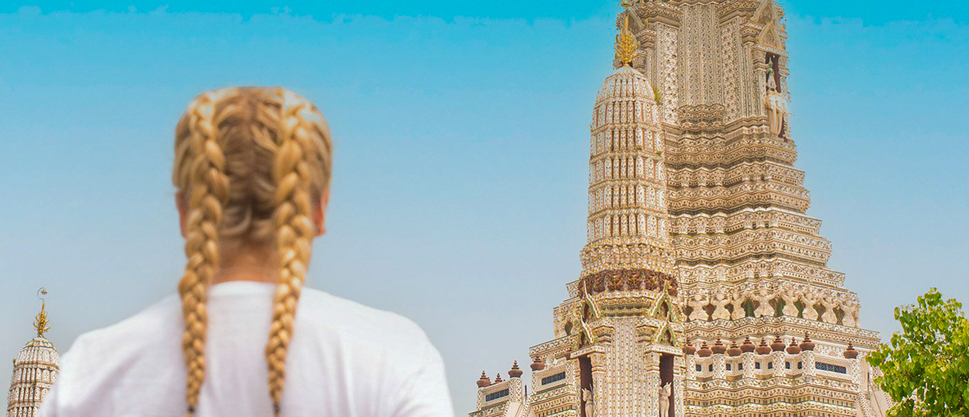 A traveler with blonde braids admires the intricate architecture of Wat Arun temple in Bangkok.