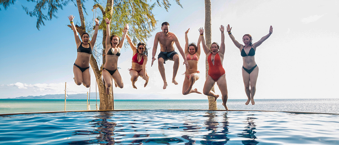 Seven happy backpackers jump high above an infinity pool overlooking the beautiful ocean in Koh Phangan.