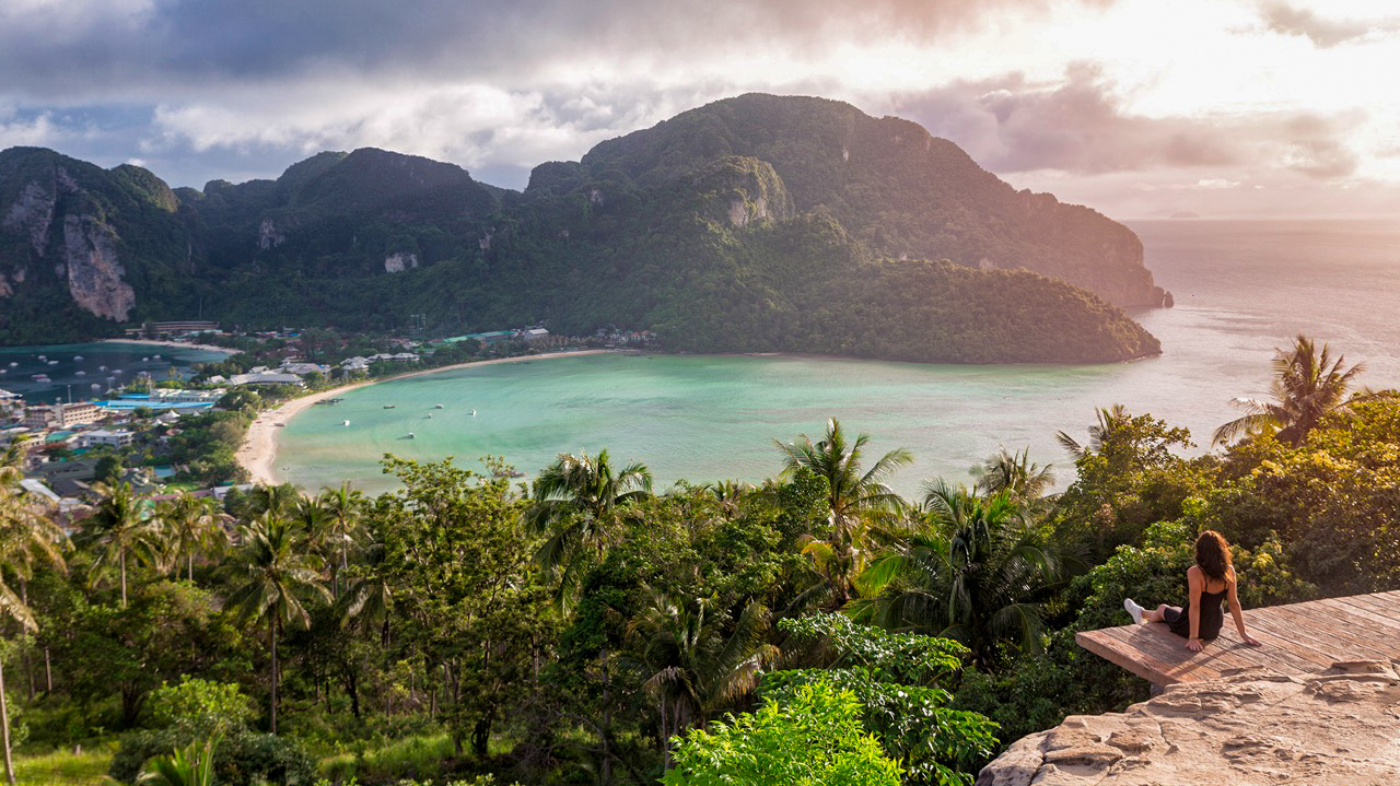 A woman sits on a viewpoint, admiring the stunning sunrise over Phi Phi Islands' turquoise bays.